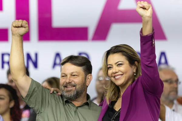 Homem de camisa cinza e mulher com blazer roxo de mãos levantadas e sorrindo