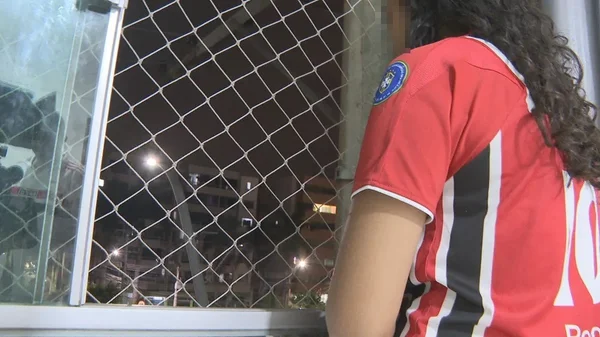 Foto colorida mostra pessoa de cabelos longos, cacheados, em frente a janela com tela de proteção, à noite; a pessoa está de costas para a câmera, vestida com camisa de time e tem o rosto borrado