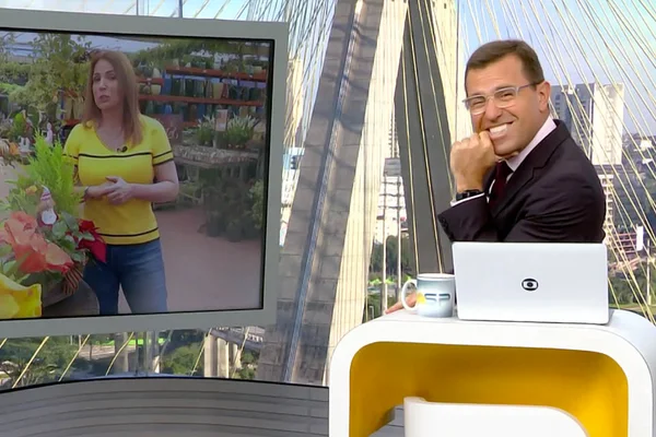 Rodrigo Bocardi sorri para a câmera enquanto Ananda Apple aparece no telão do Bom Dia SP - Metrópoles