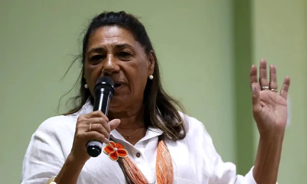 imagem colorida mostra mulher negra segurando microfone, ele veste uma blusa branca, um colar colorido com uma flor vermelha e tem a mão esquerda levantada enquanto fala. metrópoles