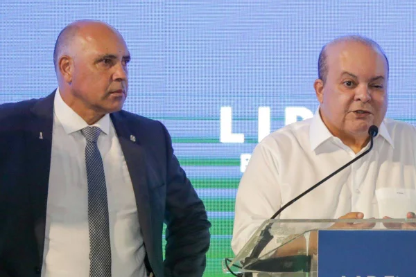 Foto colorida de Wellington Luiz, homem de terno azul, gravata azul e branca e camisa branca ao lado de Ibaneis Rocha, de camisa branca, falando ao microfone em púlpito
