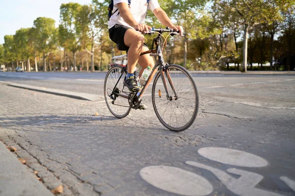 pernas de um homem em vista lateral andando de bicicleta em uma avenida em paris carregando uma garrafa de água, carro ao fundo. Metrópoles
