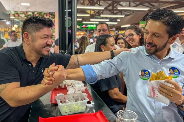 Imagem colorida mostra um comerciante de roupas pretas, em uma loja de pasteis do mercado municipal do Tucuruvi, dando a mão a Ricardo Nunes - Metrópoles