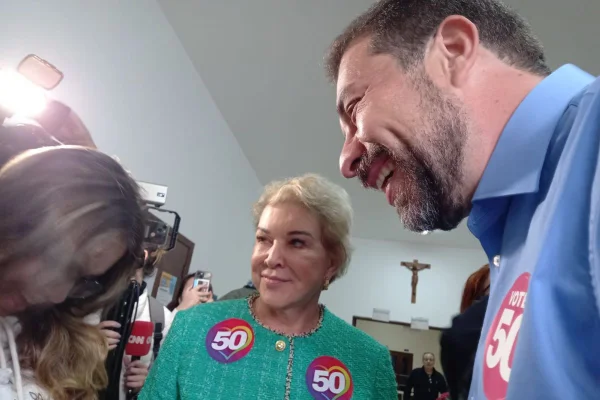 Imagem colorida mostra, no meio, mulher loira de cabelos curtos. Ela veste casaco verde claro com dois adesivos com o número 50. No seu lado esquerdo está um homem branco com cabelos escuros e barba fina, do outro lado aparece cabelo castanho claro de uma mulher - Metrópoles