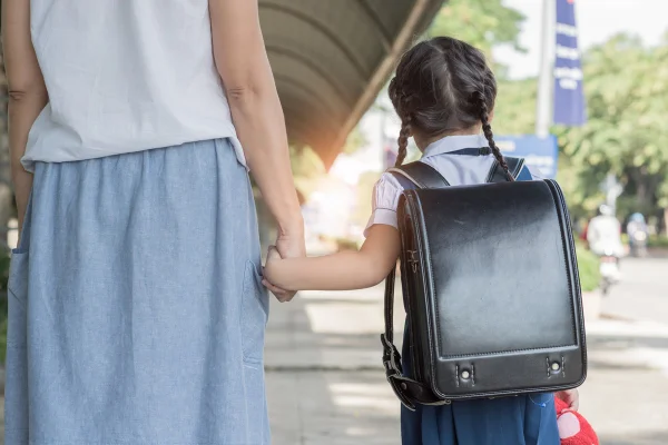 Duas moças posam para a foto. De mãos dadas à uma mulher adulta, a jovem menina carrega sua mochila preta.