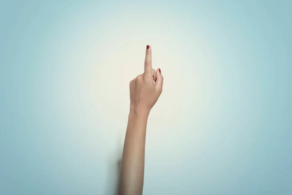 Foto colorida do braço de uma mulher com o dedo apontado para cima e as unhas vermelhas, em um fundo azul claro - Metrópoles