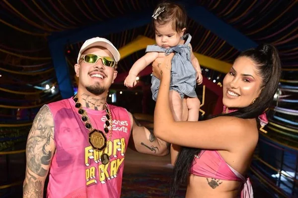 foto colorida homem com bone e oculos escuros ao lado de mulher de cabelos longos escuros segurando bebê - metrópoles