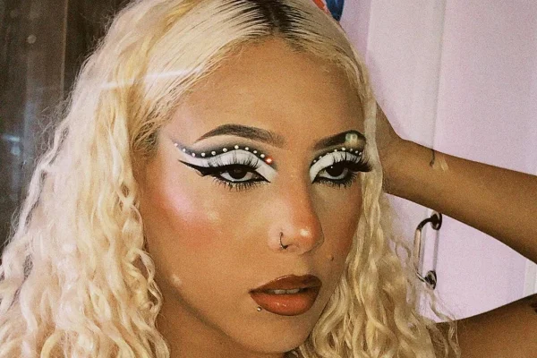 Foto colorida de mulher com cabelo cacheado e platinado. Ela está com uma maquiagem de delineado nos olhos - Metrópoles
