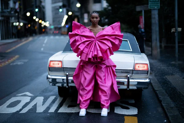 Modelo com look rosa volumoso, em frente a carro, na campanha da marca Juliana Jabour