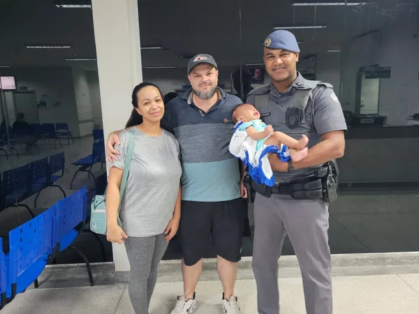 Imagem colorida mostra um policial militar (PM) segurando um bebê no colo ao lado dos pais da criança; o PM salvou a vida do menino, que se engasgou com leite materno - Metrópoles