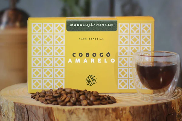 Caixa de café apoiada em uma tora de madeira com grãos e uma xícara de café