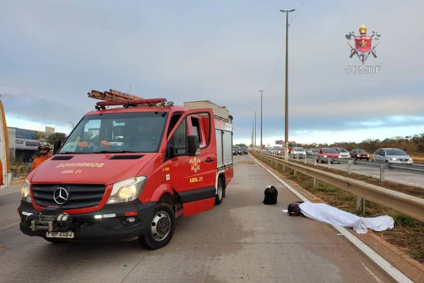 Foto colorida de corpo coberto por lençol no chão e ambulância do CBMDF