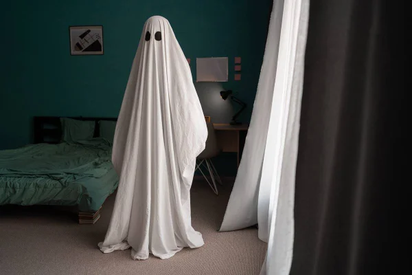 Foto colorida de uma pessoa coberta com um lençol branco imitando um fantasma em frente a uma cama desarrumada - Metrópoles