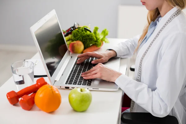 Foto mostra uma nutricionista em frente ao computador trabalhando. Na mesa tem algumas frutas e vegetais. Metrópoles