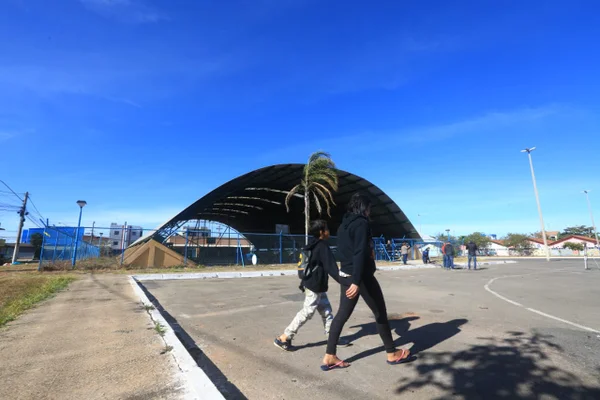 duas pessoas andando em frente a uma quadra de esportes que é um grande galpão azul