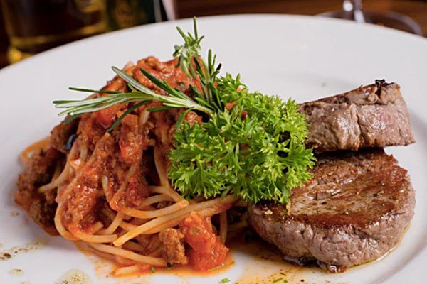 Na foto temos um prato com macarrão com molho vermelho e dois pedaços de carne ao lado