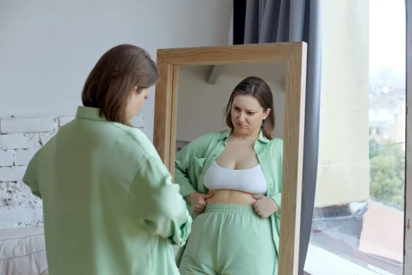 Foto colorida de mullher branca apertando a barriga, vestida com blusa e shorte verde em frente o espelho -Metrópoles.