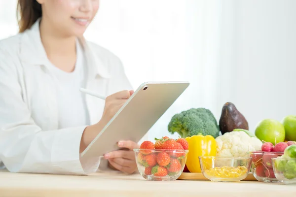 Foto colorida de mulher com jaleco e estetoscópio no pescoço. Ela está com um tablet na mão e ao lado tem potes de vidro com frutas e legumes - Metrópoles