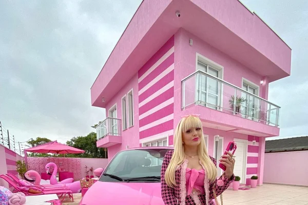 Foto colorida de mulher branca e loira, sentada próximo a um carro pink. Ao fundo, tem uma casa pintada de rosa - Metrópoles