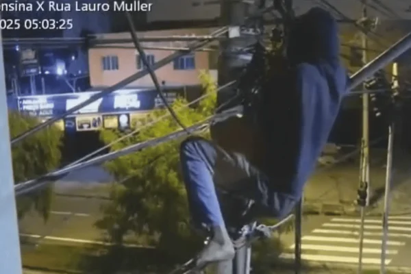 Imagem colorida de um homem encapuzado pendurado em um poste cortando fios - Metrópoles