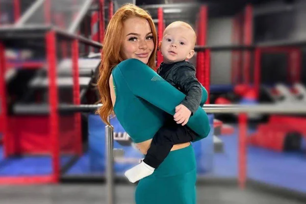 Mulher ruiva, jovem, vestindo roupa verde, segurando um bebê no colo com fundo embaçado - Metrópoles