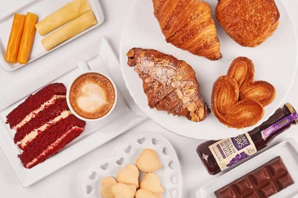Na foto temos um fundo branco comum prato com queijos, um prato com red velvet e um café, um prato com biscoitos, um prato com chocolate, uma garrafa de suco de uva e um prato com croissants