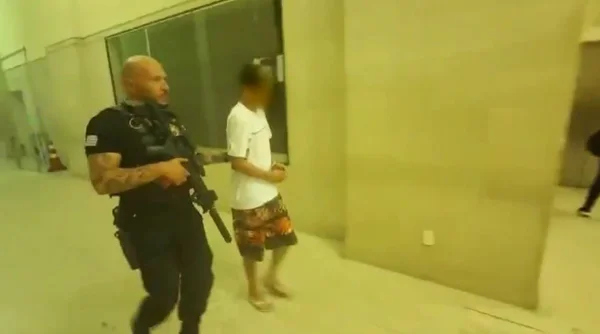 Imagem de policial conduzindo homem de camiseta branca e bermuda, em corredor - Metrópoles