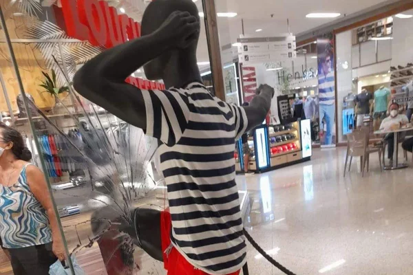 Manequim preto com blusa listrada e bermuda vermelha quebrando a vitrine de vidro da marca Reserva, em corredor de shopping