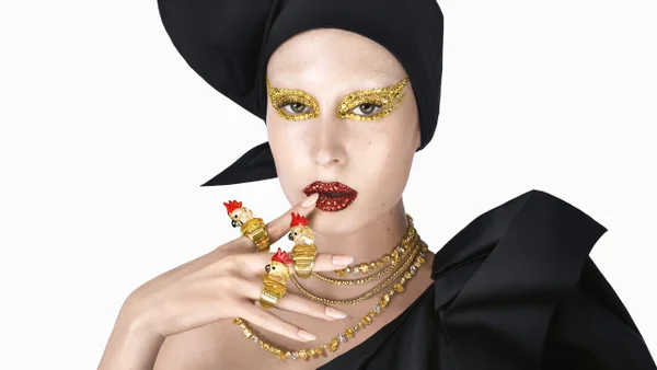 Na imagem com cor, uma mulher branca em um fundo branco usando um chapéu e vestido preto com detalhes dourado nos olhos - Metrópoles