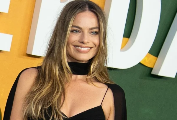 Foto colorida da atriz Margot Robbie sorrindo com um vestido preto e o cabelo solto - Metrópoles