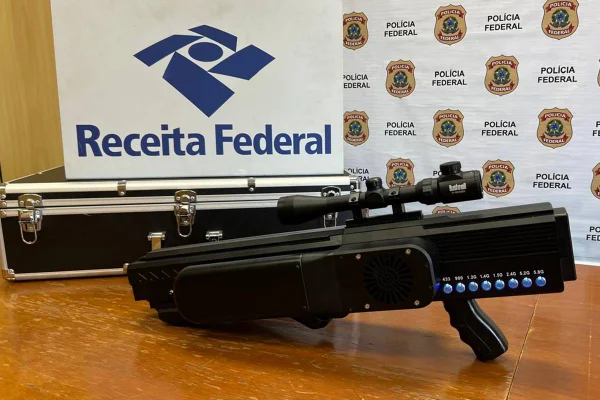 Foto colorida de fuzil anti-drones em cima de mesa com símbolos da Receita Federal e Polícia Federal