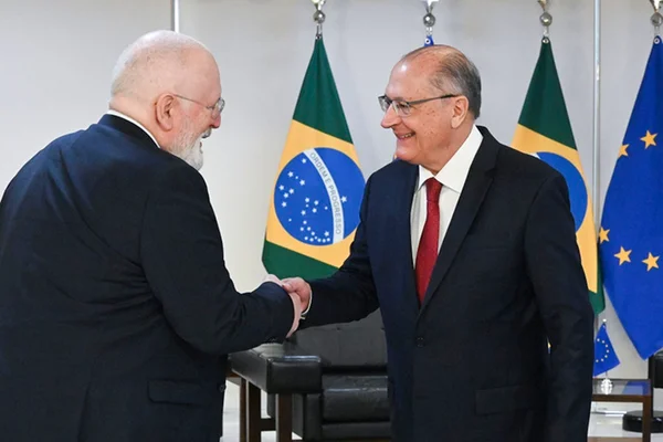 Frans Timmermans, representante da União Europeia, e Geraldo Alckmin, presidente em exercício do Brasil, apertam as mãos após encontro. Eles sorriem e se vê a fundo as bandeiras respectivas - Metrópoles