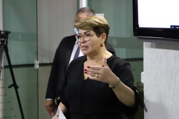 Mulher loira e branca vestida de preto com uma expressão de fala e uma das mãos levantadas