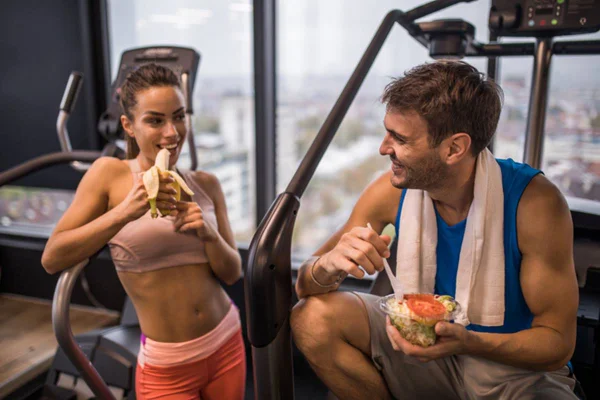 Foto colorida: casal na academia comendo frutas e saladas