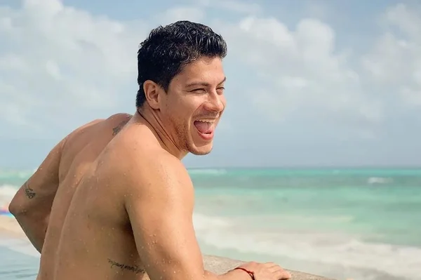 Na imagem colorida, um homem está posicionado no centro. Ele tem está sem camisa, tem cabelos curtos e pretos, tem barba curta e está com fisionomia seria