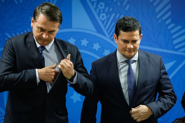 Na imagem colorida, dois homens estão centralizados. Eles usam terno e gravada escuro, possuem cabelo curto e ambos são brancos
