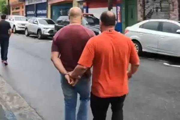Em foto colorida policial de camiseta laranja, de costas, conduz homem careca de camiseta vinho à frente, de costas também - Metrópoles