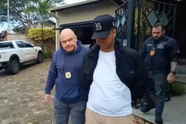 Policial branco e careca, usando jaqueta de moletom azul escura e calça jeans conduz homem pardo com boné preto, cabeça baixa, jaqueta preta, camiseta branca e calça creme - Metrópoles