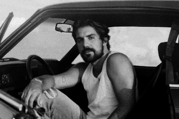 O cantor Gabriel Gonti lança seu novo single. Na foto preta e branca, ele está dentro de carro antigo, com as pernas para fora e segura um óculos escuros. Ele é branco, tem cabelo liso e preto e barba - Metrópoles