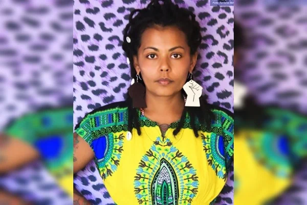 Mulher negra com roupa amarela estampada em frente a parede roxa com estampa de onça