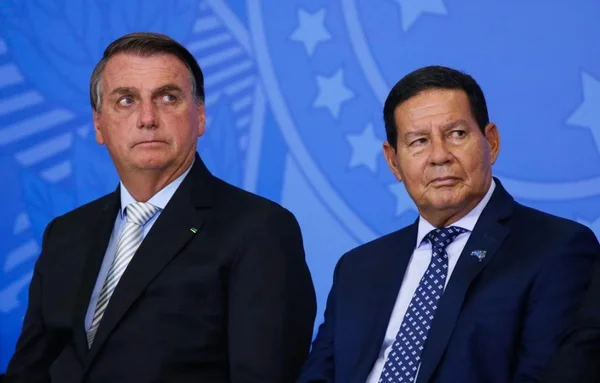Na imagem colorida, dois homens estão posicionados no centro. Eles usam terno e gravata na cor preta e camiseta branca. Eles tem cabelos curtos e escuros
