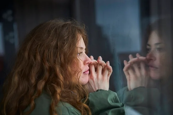 Mulher branca de cabelos ruivos e cacheados está olhando pensativa pela janela, que reflete o seu olhar triste. A imagem representa uma pessoa com depressão - Metrópoles