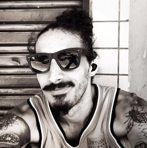 Foto em preto e branco. Homem de bigode e cavanhaque veste camisa regata. Também usa óculos escuros e possui cabelo preso