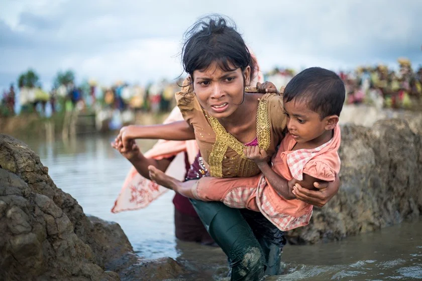 Uma mãe Rohingya atravessa a água para chegar à fronteira de
Myanmar (Birmânia) a Bangladesh, perto do vilarejo de Anzuman Para em Palong Khali. Estimados 500 mil Rohingya buscaram refúgio em Bangladesh entre o final de agosto e outubro de 2017. A ACNUR colaborou com as autoridades para criar um centro de trânsito para se prepararem para um fluxo maior de pessoas, já que cerca de 11 mil pessoas passaram pela fronteira em um dia. Eles atravessaram a pé através de diversos pontos, e muitos vieram da área Buthidaung no estado de Rakhine, no norte do Myanmar. Alguns afirmam fugir de matança e ateamento de fogo. Outros disseram que fugiram por medo da violência que antecipavam. Para chegar a Bangladesh, tiveram de andar durante dias, muitos carregando crianças. Atravessaram pântanos antes de nadar através do rio Naf que divide os dois países. A ACNUR tenta acomodar a maior quantidade de pessoas possíveis nos acampamentos em Kutupalong e Balukhali, fornecendo itens de emergência Uma mãe Rohingya atravessa a água para chegar à fronteira de
Myanmar (Birmânia) a Bangladesh, perto do vilarejo de Anzuman Para em Palong Khali. Estimados 500 mil Rohingya buscaram refúgio em Bangladesh entre o final de agosto e outubro de 2017. A ACNUR colaborou com as autoridades para criar um centro de trânsito para se prepararem para um fluxo maior de pessoas, já que cerca de 11 mil pessoas passaram pela fronteira em um dia. Eles atravessaram a pé através de diversos pontos, e muitos vieram da área Buthidaung no estado de Rakhine, no norte do Myanmar. Alguns afirmam fugir de matança e ateamento de fogo. Outros disseram que fugiram por medo da violência que antecipavam. Para chegar a Bangladesh, tiveram de andar durante dias, muitos carregando crianças. Atravessaram pântanos antes de nadar através do rio Naf que divide os dois países. A ACNUR tenta acomodar a maior quantidade de pessoas possíveis nos acampamentos em Kutupalong e Balukhali, fornecendo itens de emergência