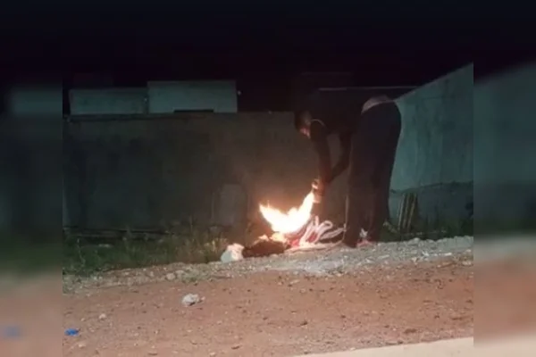 Homem abaixado ateando fogo em roupas no chão de barro à noite em frente a muro