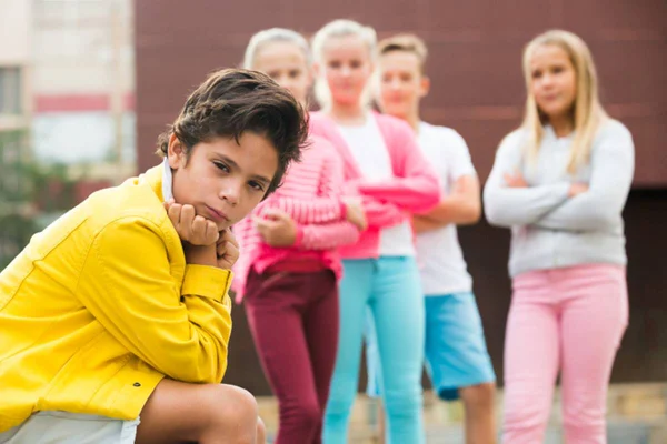 Imagem colorida de menina vestido de amarelo sentado sozinho em um banco com semblante chateado ignorando seus amigos ao fundo da imagem - Metrópoles