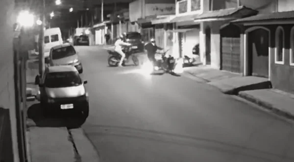 Imagem preta e branca de dois motociclistas em pé apontando armas de fogo para homem caído ao chão - Metrópoles