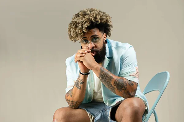 Foto de um homem negro com cabelo preto com pontas loiras. Ele usa óculos de grau, está sentado, e tem barba preta. Usa look azul com jeans - Metrópoles