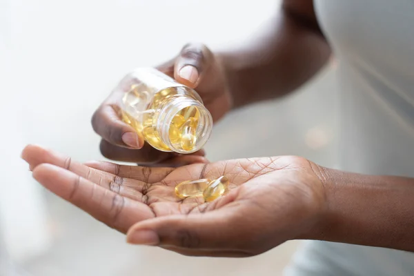 Foto mostra mão de pessoa negra com um vidro transparente pegando cápsulas de suplementos amareladas - Metrópoles