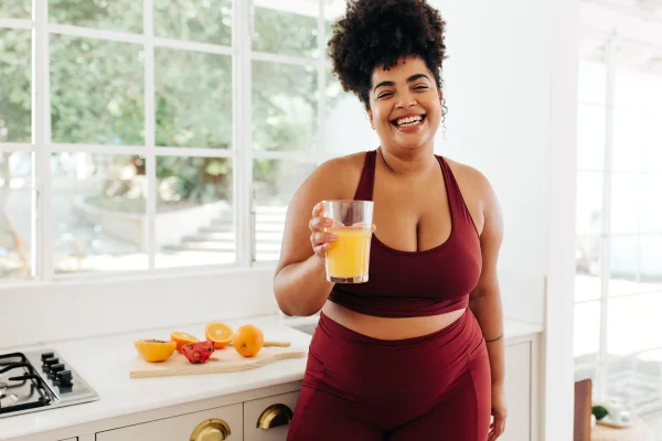 Foto colorida de mulher, com roupa de ginástica, tomando suco de laranja - Metrópoles